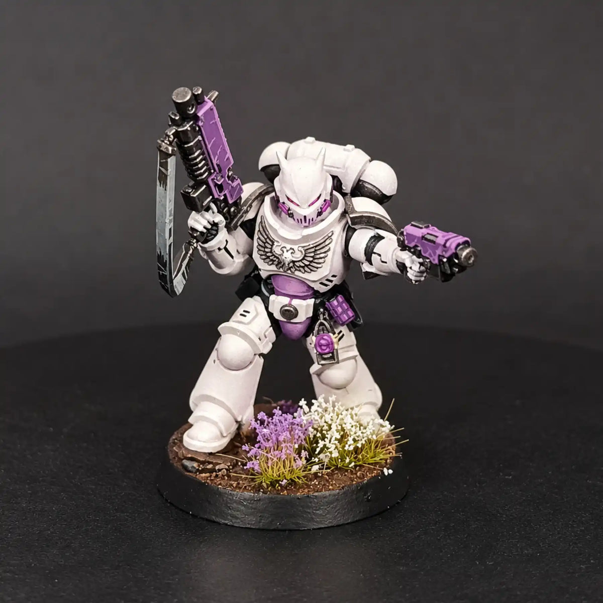 A painted sci-fi miniature figurine of a white armored warrior holding a purple and silver sword in one hand and a purple gun in the other, standing on a circular base with small purple and white flowers against a dark gray background.