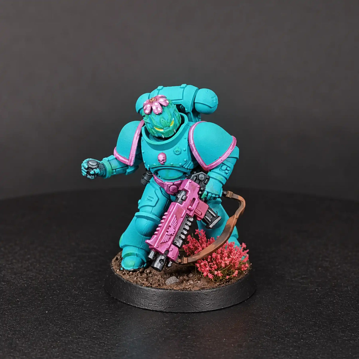 A painted fantasy miniature figurine of a kneeling armored warrior in bright turquoise and pink, holding a pink weapon, mounted on a round black base with small red flowers and dark soil, photographed against a dark gray studio background.